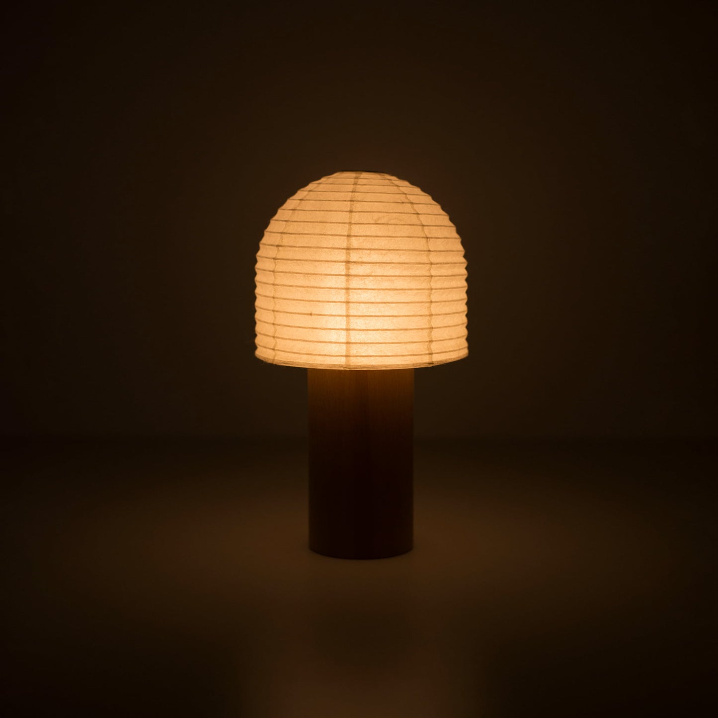 A portable paper lamp with a wooden handle and bell-shaped cylindrical paper top emitting warm light in a dark room