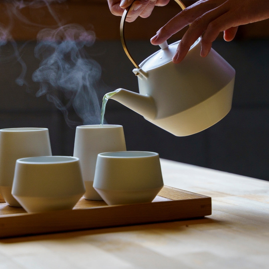Hand pouring hot Japanese green tea into yunomi cups from a Japanese teapot set