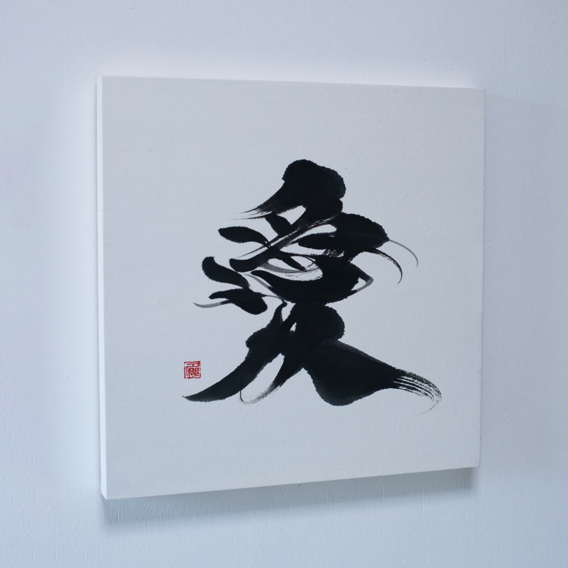 A wall art panel displaying '愛' (love) in Japanese calligraphy, mounted on a white wall. 
