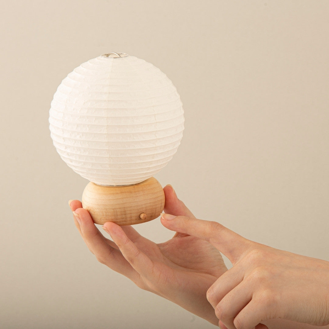 A hand holding the Japanese paper table lamp 'Mini Circle' while pointing at the switch.