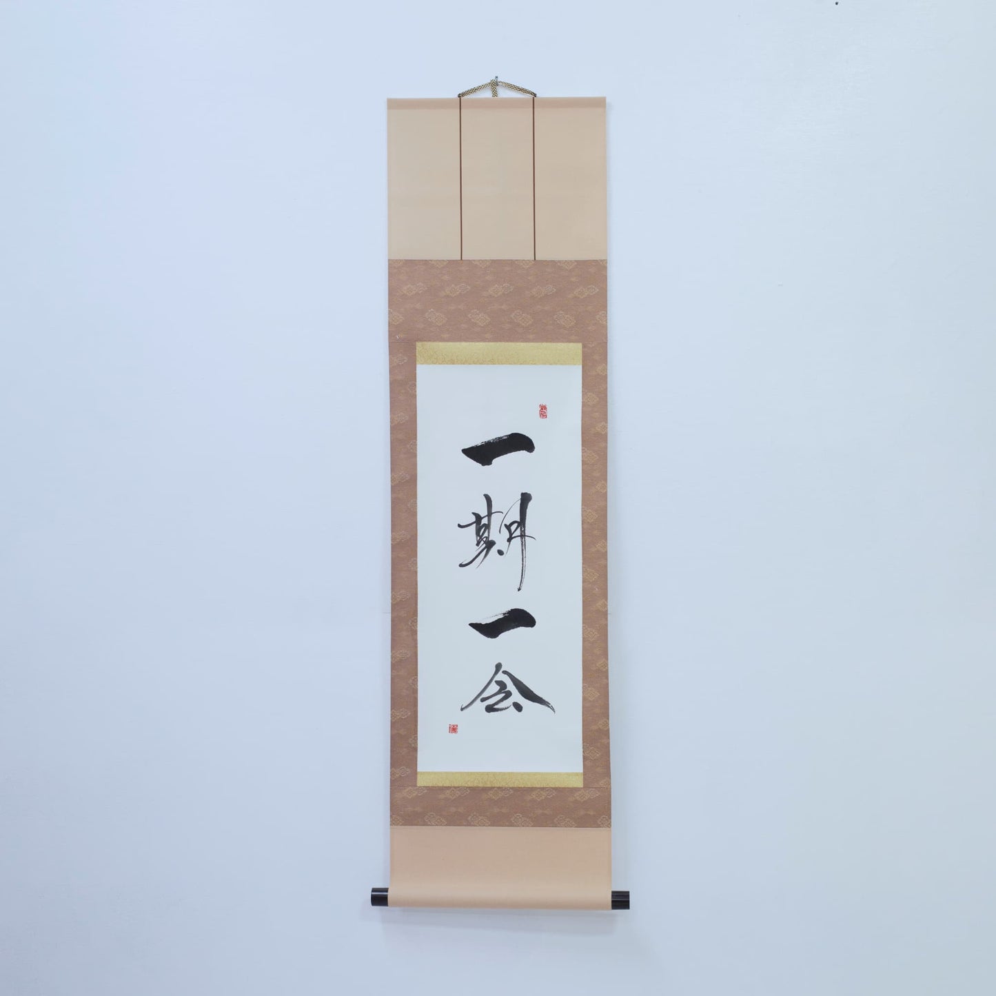 Brown hanging scroll with Japanese calligraphy of '一期一会' (one-time, one meeting) on white rice paper, mounted on a white wall