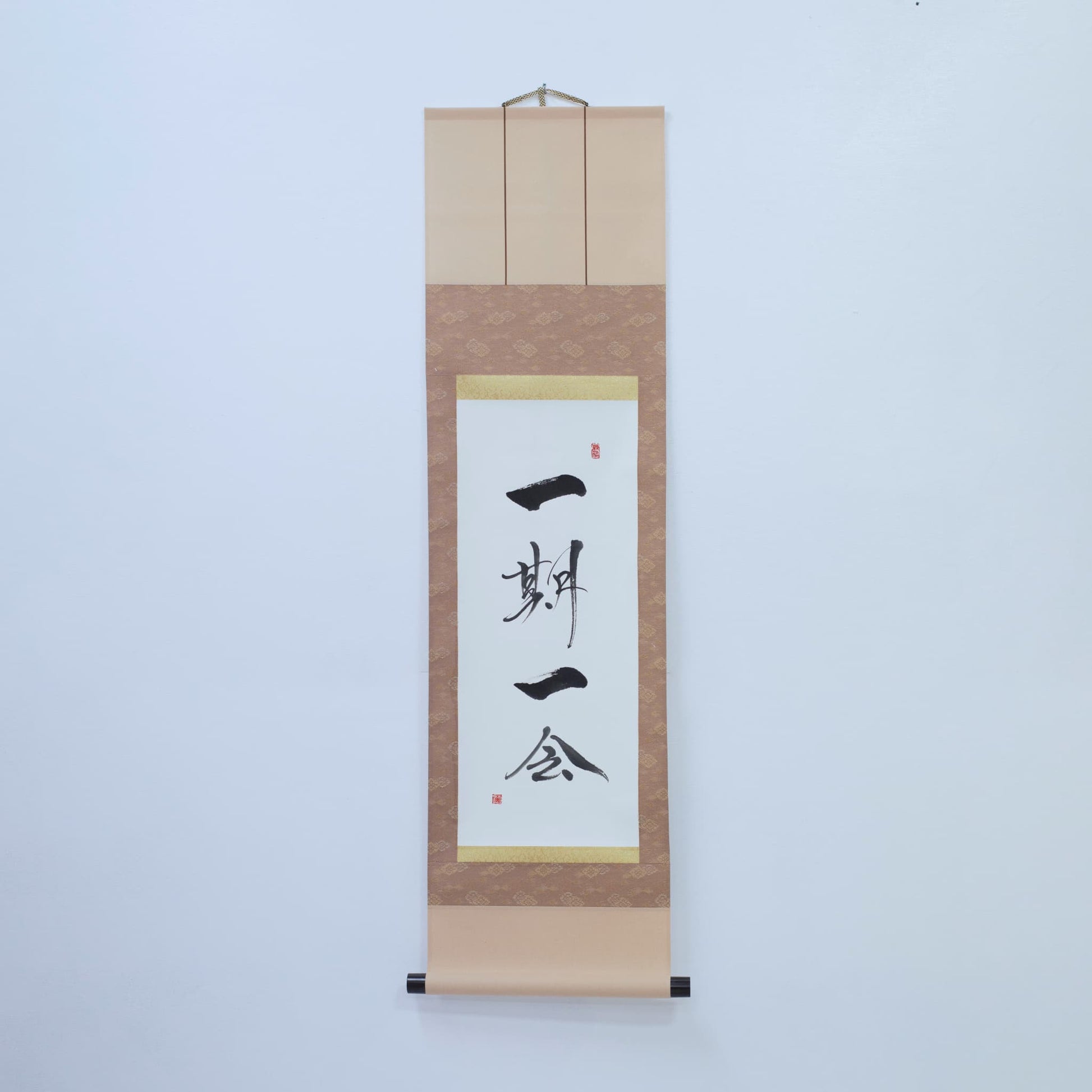 Brown hanging scroll with Japanese calligraphy of '一期一会' (one-time, one meeting) on white rice paper, mounted on a white wall