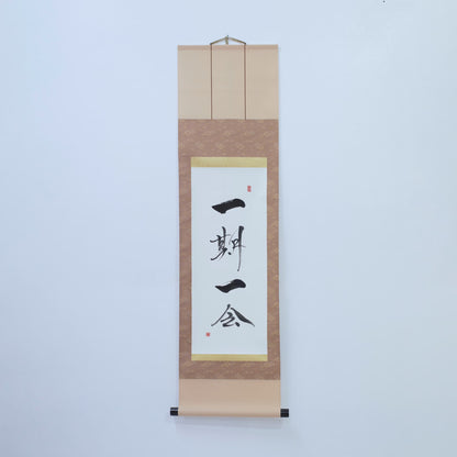 Brown hanging scroll with Japanese calligraphy of '一期一会' (one-time, one meeting) on white rice paper, mounted on a white wall
