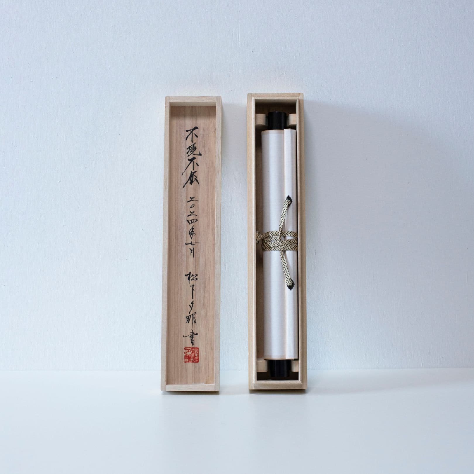 A long, slender wooden box with its lid, used for storing a traditional Japanese hanging scroll, showing the scroll rolled up inside