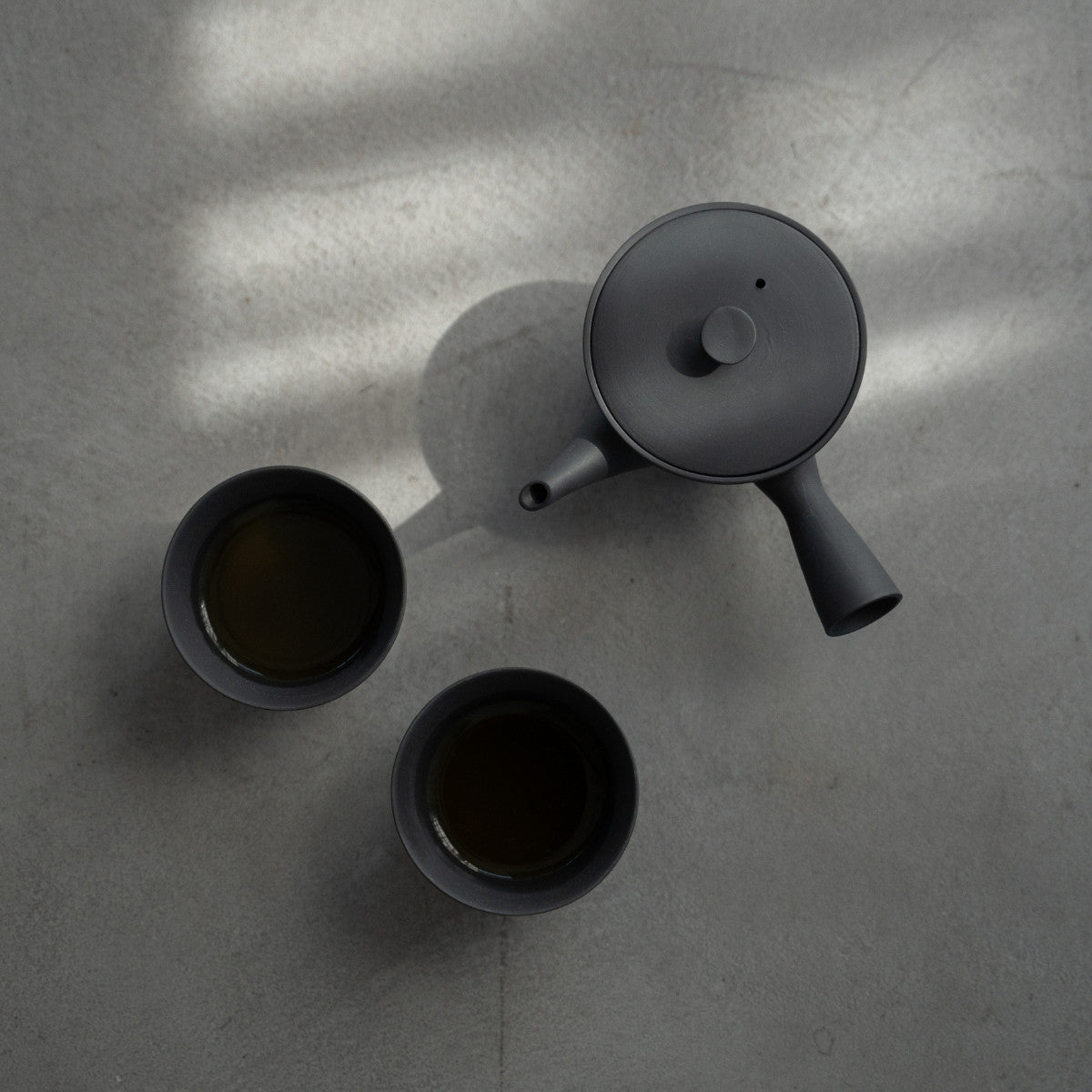 Black kyusu "Sencha" and filled Japanese tea cups on a concrete surface for minimalists enjoying a quiet tea break.