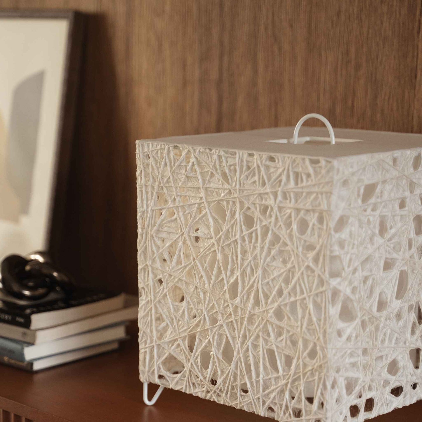 White cube shaped Japanese paper lamp Me Look 03 sitting unlit on a wooden console table near a bed background.