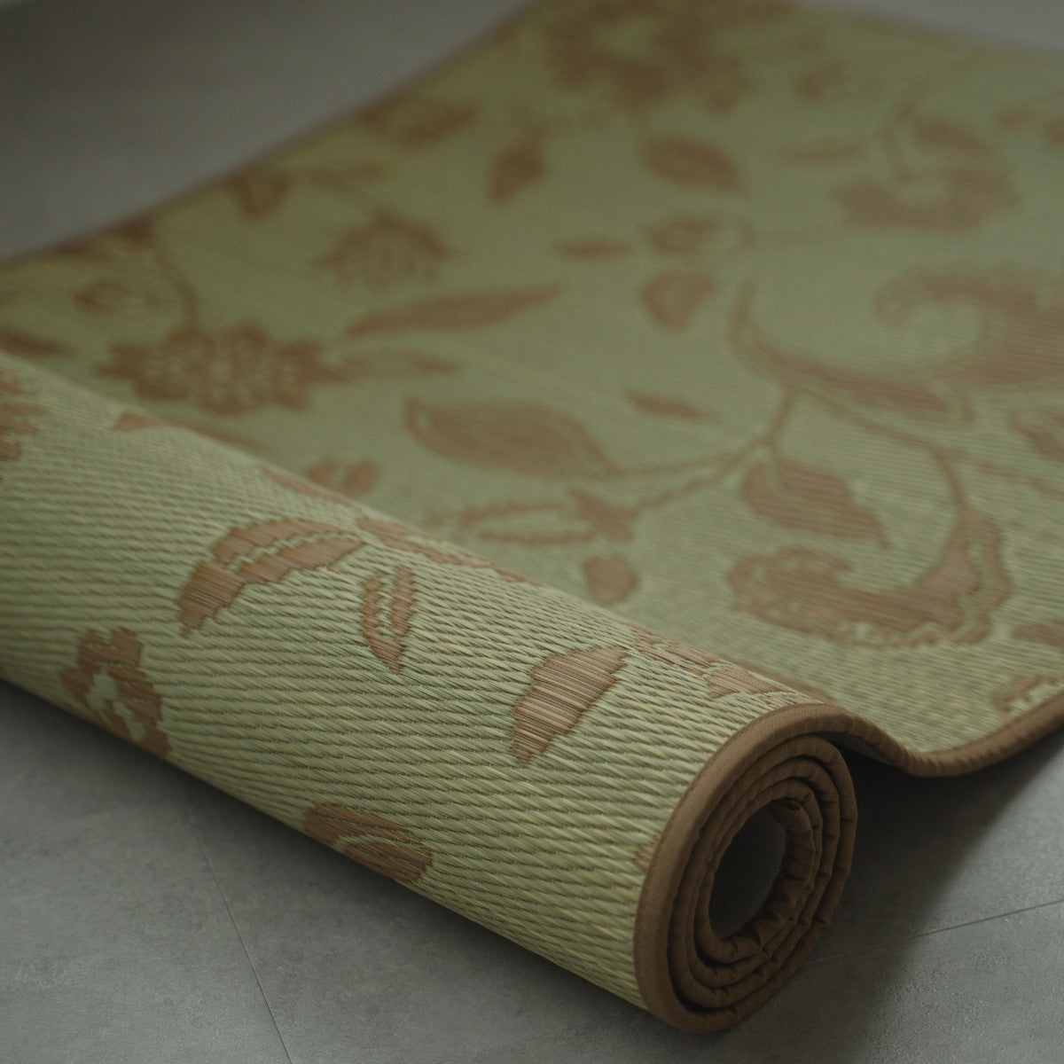 Close-up van Japanse Tatami Yoga Mat "Sarasa" Beige, gedeeltelijk opgerold op een grijze vloer, met zichtbare geweven textuur van natuurlijk rushgras.