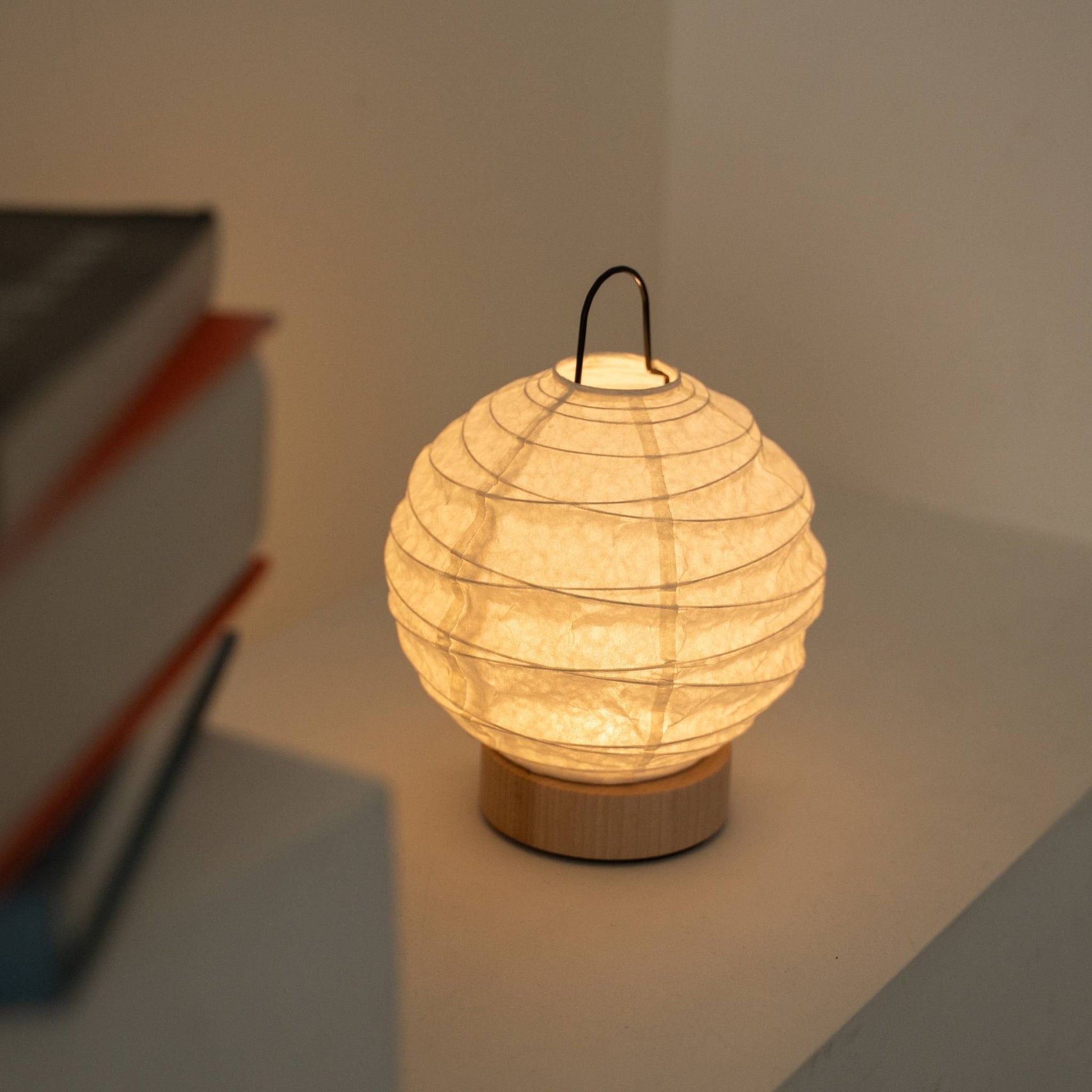 Glowing round Japanese paper table lamp "Yuhi" serving as a cozy accent lamp next to books on a white console table.