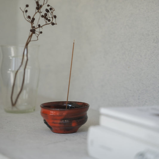 Crimson red Japanese incense burner with a lit stick on a light shelf, safely catching ash for a serene morning meditation.