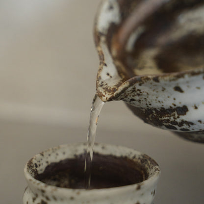 Sake fließt aus einer strukturierten japanischen Zui-Sakekaraffe in eine weiße Keramik-Sake-Tasse, um warmen Sake mühelos zu genießen