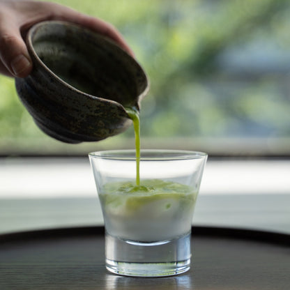 Pouring matcha from a rustic Japanese matcha bowl with spout into milk in a glass to make a latte.