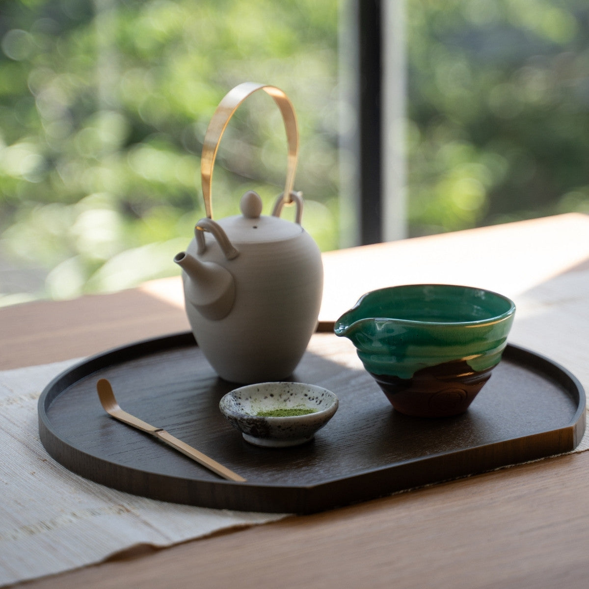Emerald Japanese matcha bowl with spout and tea tools on a dark tray by a bright window, set to make lattes for guests.