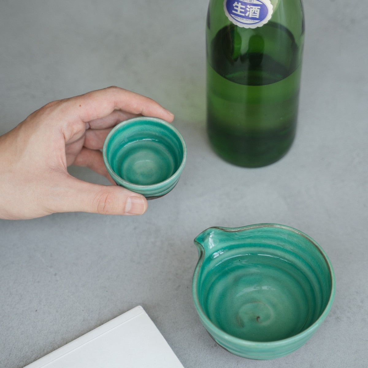 Eine Hand hält einen smaragdgrünen japanischen Sakebecher über einen grauen Tisch, mit einer passenden Sakekaraffe bereit für einen reifen Abenddrink.