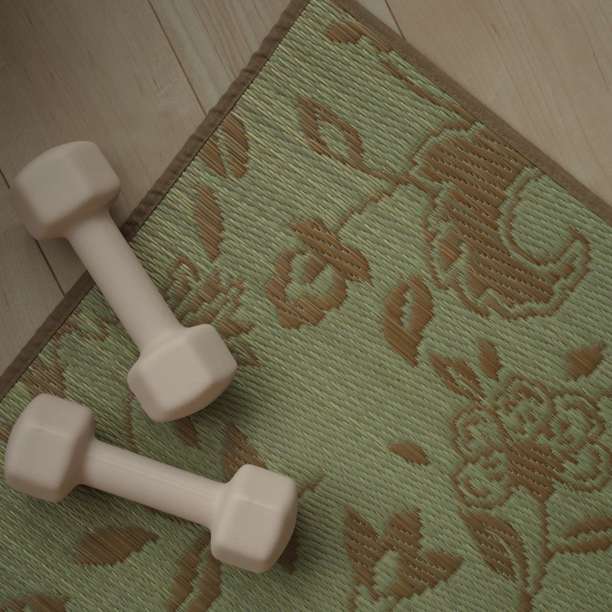 Een bovenaanzicht van een Japanse Tatami Yoga Matras "Sarasa" Beige op een houten vloer met een klein dumbbell, met de nadruk op het botanische patroon.