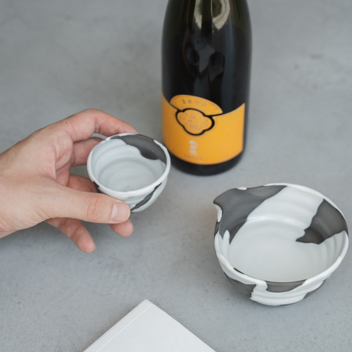 Eine Hand hält eine weiße japanische Sake-Tasse von Flow neben einer Karaffe und einem Buch auf einem grauen Tisch für ein entspannendes Sake-Ritual am Abend