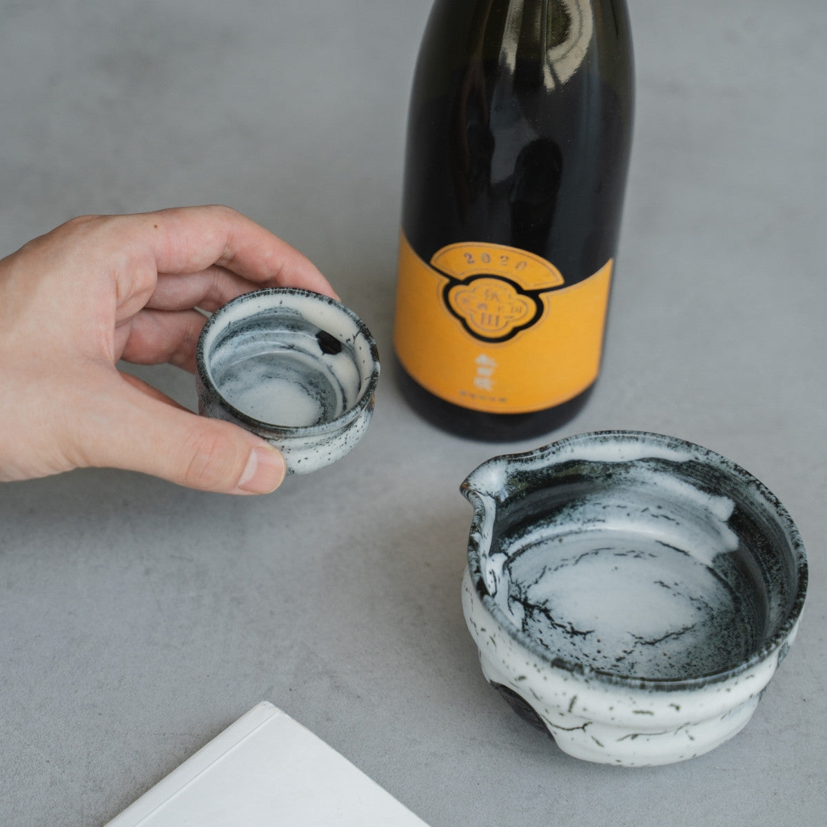 Eine Hand hält einen japanischen Sake-Becher von Creative Cloud neben einer Sake-Karaffe und einem Buch für einen entspannten Wabi-Sabi-Abend