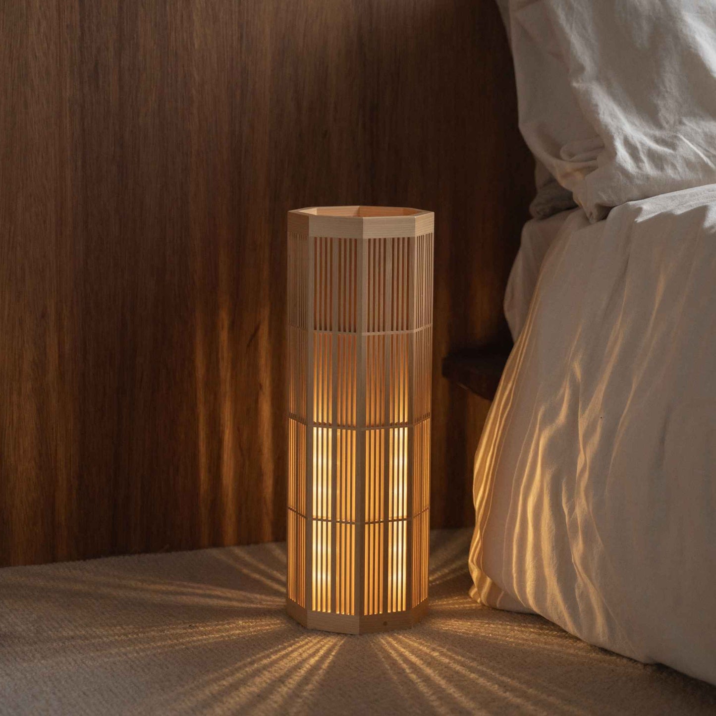 Lampe sur pied japonaise "SAN" Grand modèle en bois douillet sur le sol d'une chambre, projetant des ombres radiales apaisantes en tant que lampe d'ambiance.