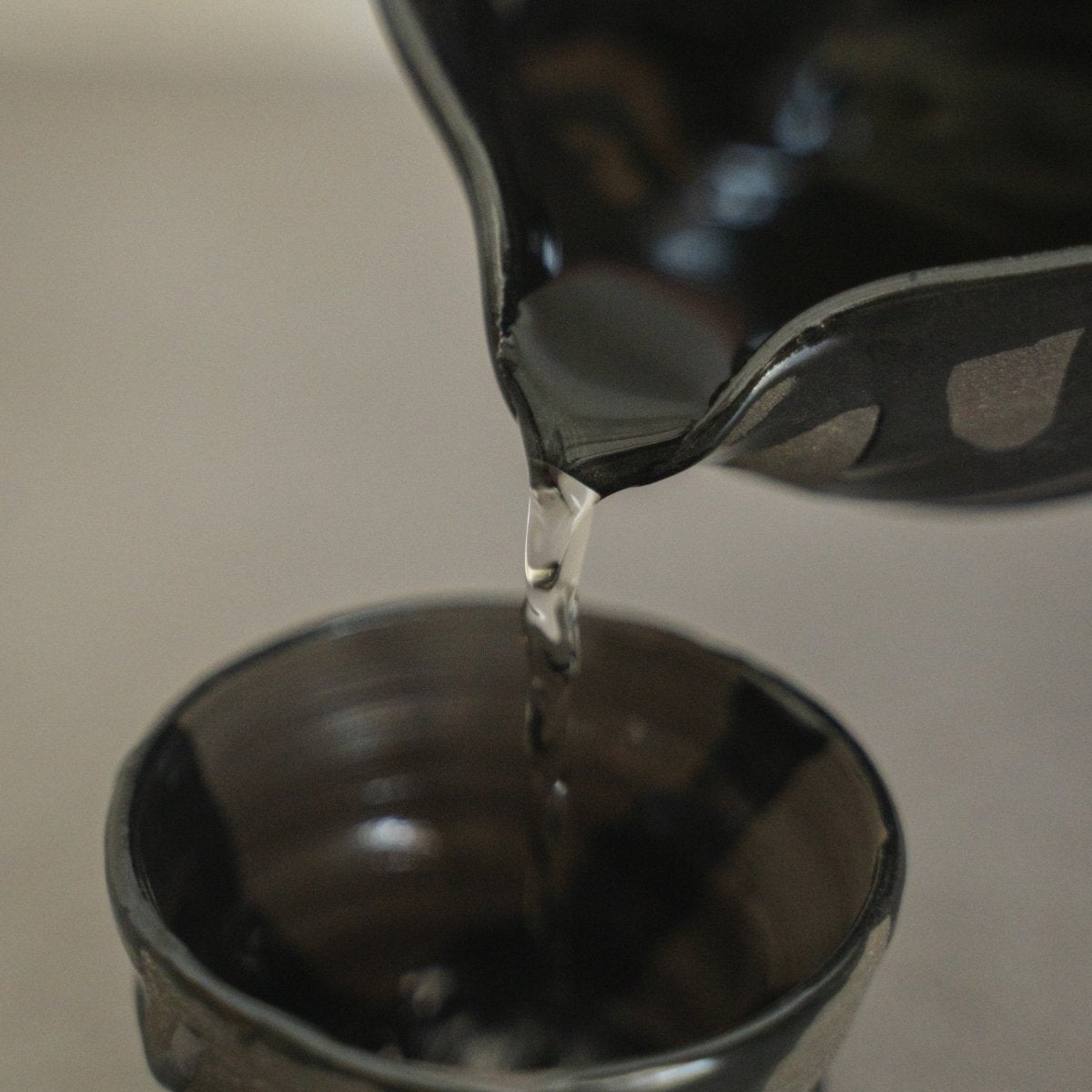 Nahaufnahme von Flüssigkeit, die aus dem Ausguss einer Sumi Black japanischen Sake-Karaffe in eine handgetöpferte Sake-Tasse fließt, für den achtsamen Genuss von heißem Sake.