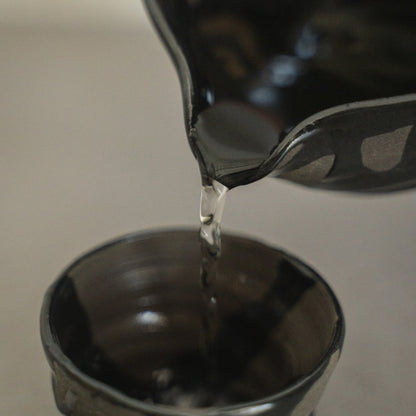 Nahaufnahme von Flüssigkeit, die aus dem Ausguss einer Sumi Black japanischen Sake-Karaffe in eine handgetöpferte Sake-Tasse fließt, für den achtsamen Genuss von heißem Sake.