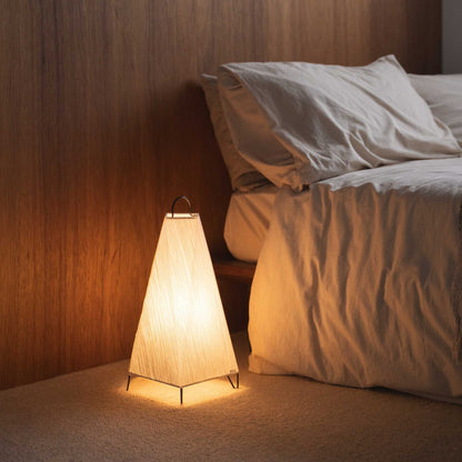 Lit Japanese paper floor lamp ORI small casting a warm glow as a cozy ambient lamp beside a bed in a relaxing bedroom.