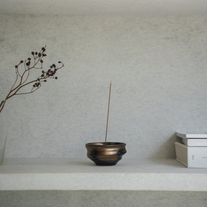 Gold and black Japanese incense burner with a lit stick on a light shelf, keeping ash contained for a calm meditation.