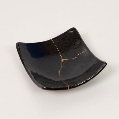 A sophisticated black square kintsugi plate rests directly on a white surface highlighting the authentic gold repair