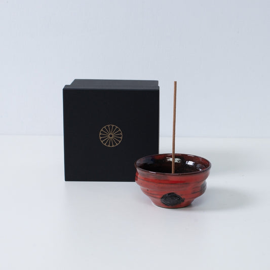 Crimson and black Japanese incense holder "Nobel Red" pottery bowl with a stick next to a black gift box with a gold chrysanthemum crest, Japan.