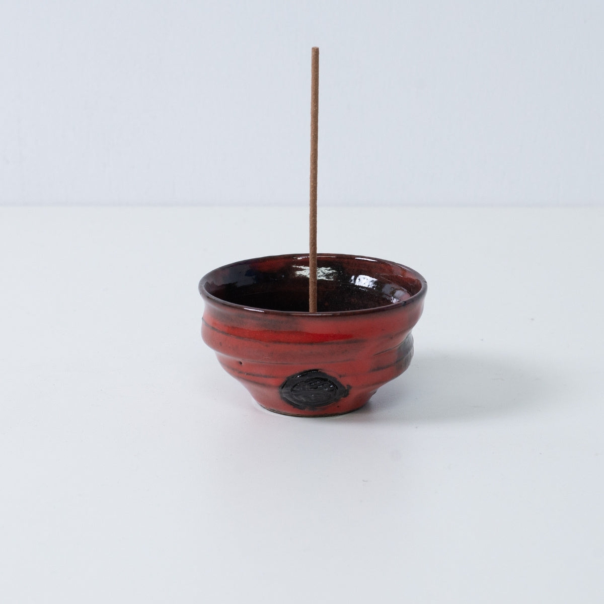 Standalone Japanese incense holder "Nobel Red" pottery bowl with a stick, showing crimson and black glaze and an artisan seal.