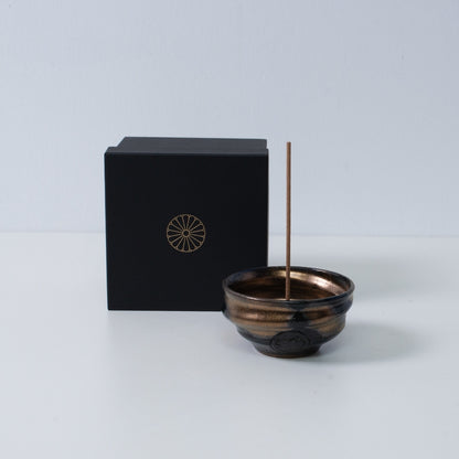 Gold and black Japanese incense holder "Zen" pottery bowl with a stick next to a black gift box with a gold chrysanthemum crest, Japan.