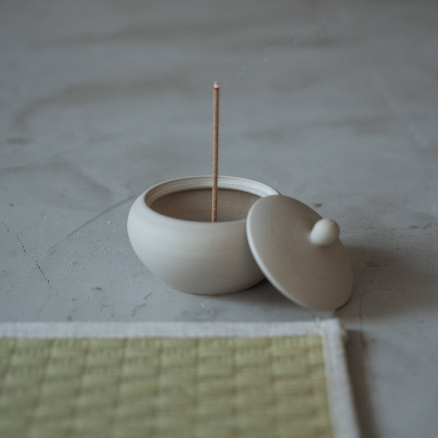 White japanese incense burner shaped like a monk bowl with a burning stick and rising smoke near a tatami mat.