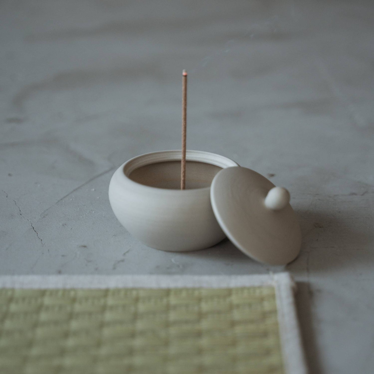 White japanese incense burner shaped like a monk bowl with a burning stick and rising smoke near a tatami mat.