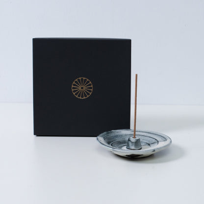 White and black Japanese incense holder "Creative Cloud" pottery with a stick next to a black gift box with a gold chrysanthemum crest.