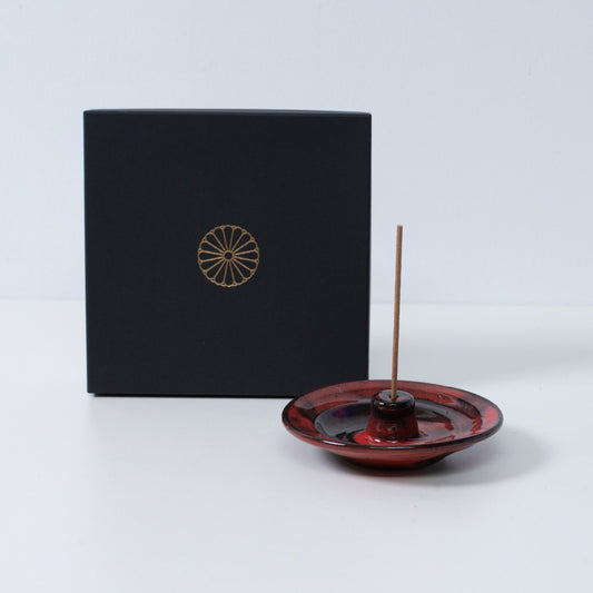 Crimson and black Japanese incense holder "Nobel Red" pottery with a stick next to a black gift box with a gold chrysanthemum crest, made in Japan.