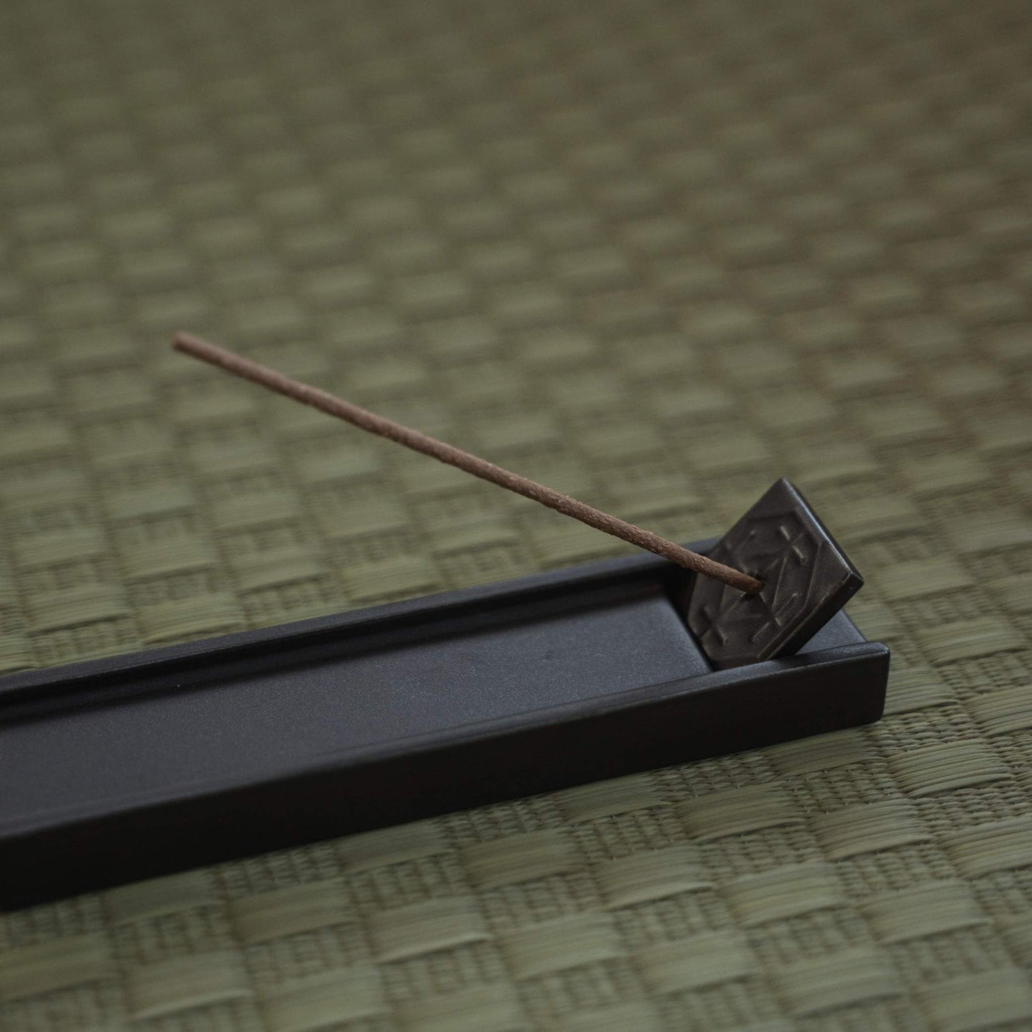 Rectangular dark japanese incense holder on a tatami mat with a burning stick creating a calm atmosphere.