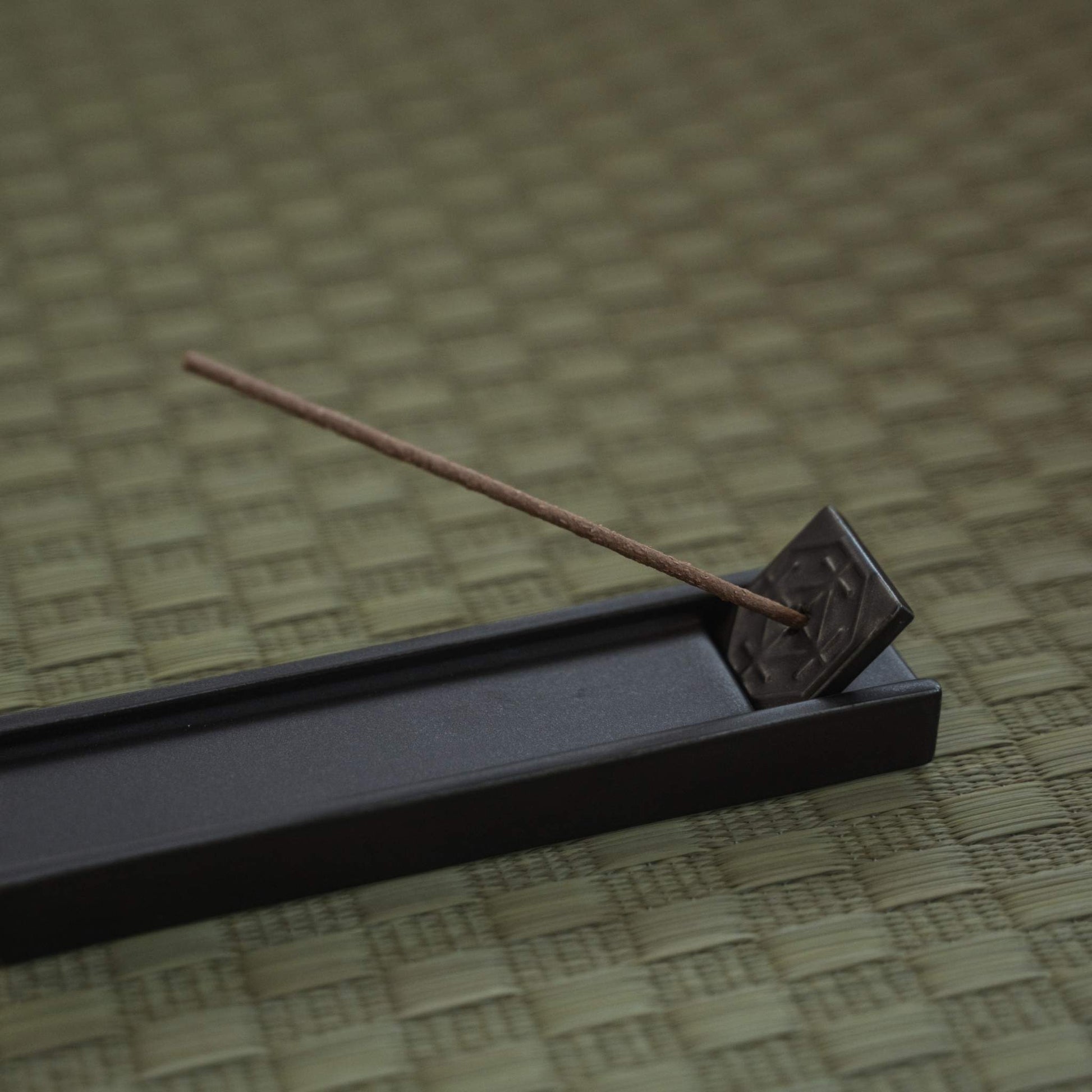 Rectangular dark japanese incense holder on a tatami mat with a burning stick creating a calm atmosphere.