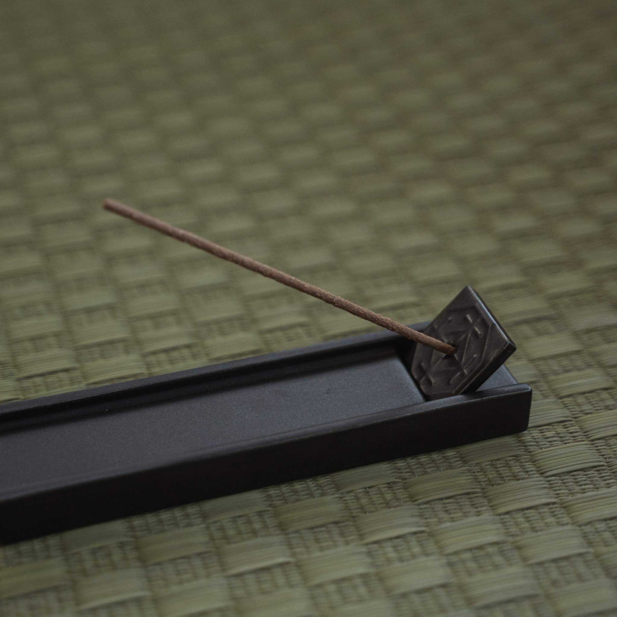 Rectangular dark japanese incense holder on a tatami mat with a burning stick creating a calm atmosphere.