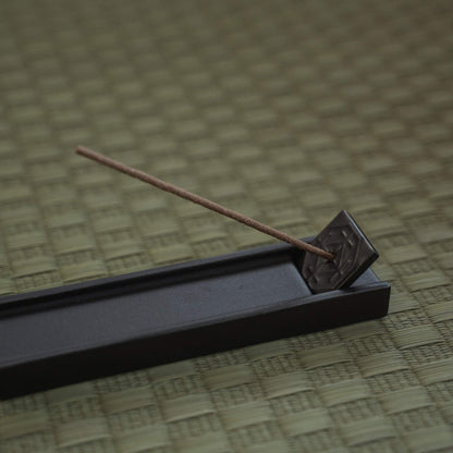 Rectangular dark japanese incense holder on a tatami mat with a burning stick creating a calm atmosphere.