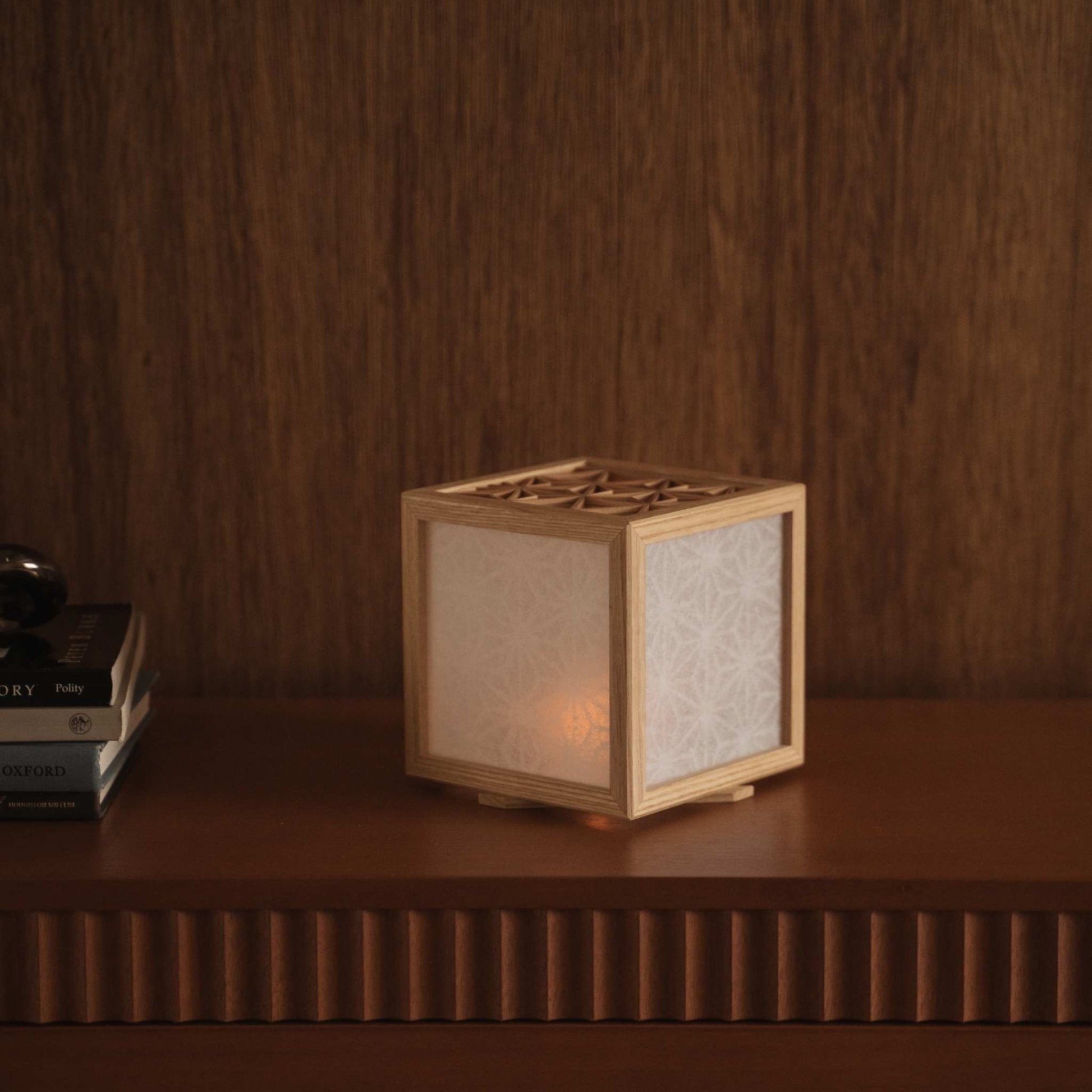 Glowing japanese kumiko lamp cover sitting on a dark wooden shelf next to books creating a warm and elegant atmosphere.