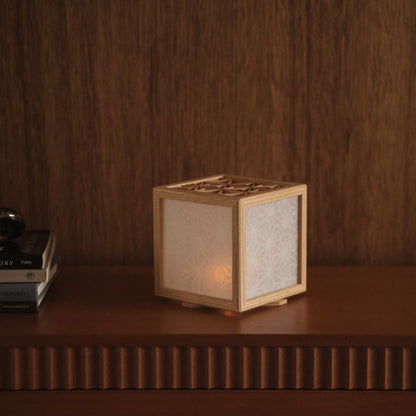 Glowing japanese kumiko lamp cover sitting on a dark wooden shelf next to books creating a warm and elegant atmosphere.