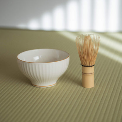 Side view of a ribbed white porcelain matcha bowl sitting on a traditional tatami mat next to a tea whisk.