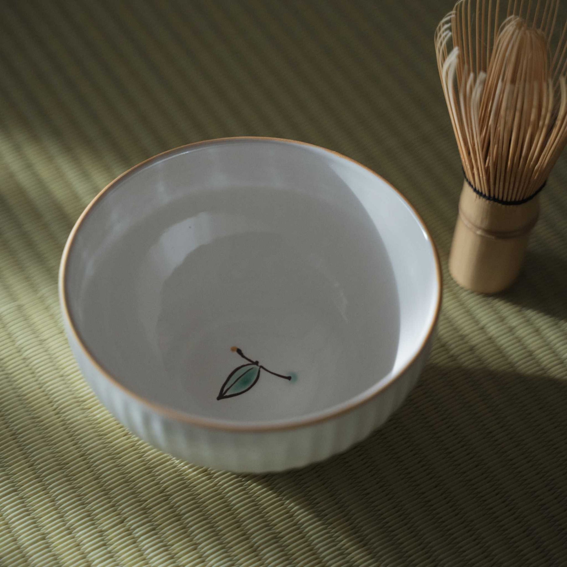 Interior view of a white japanese matcha bowl with a small painted bud next to a 
bamboo chasen on a tatami mat.