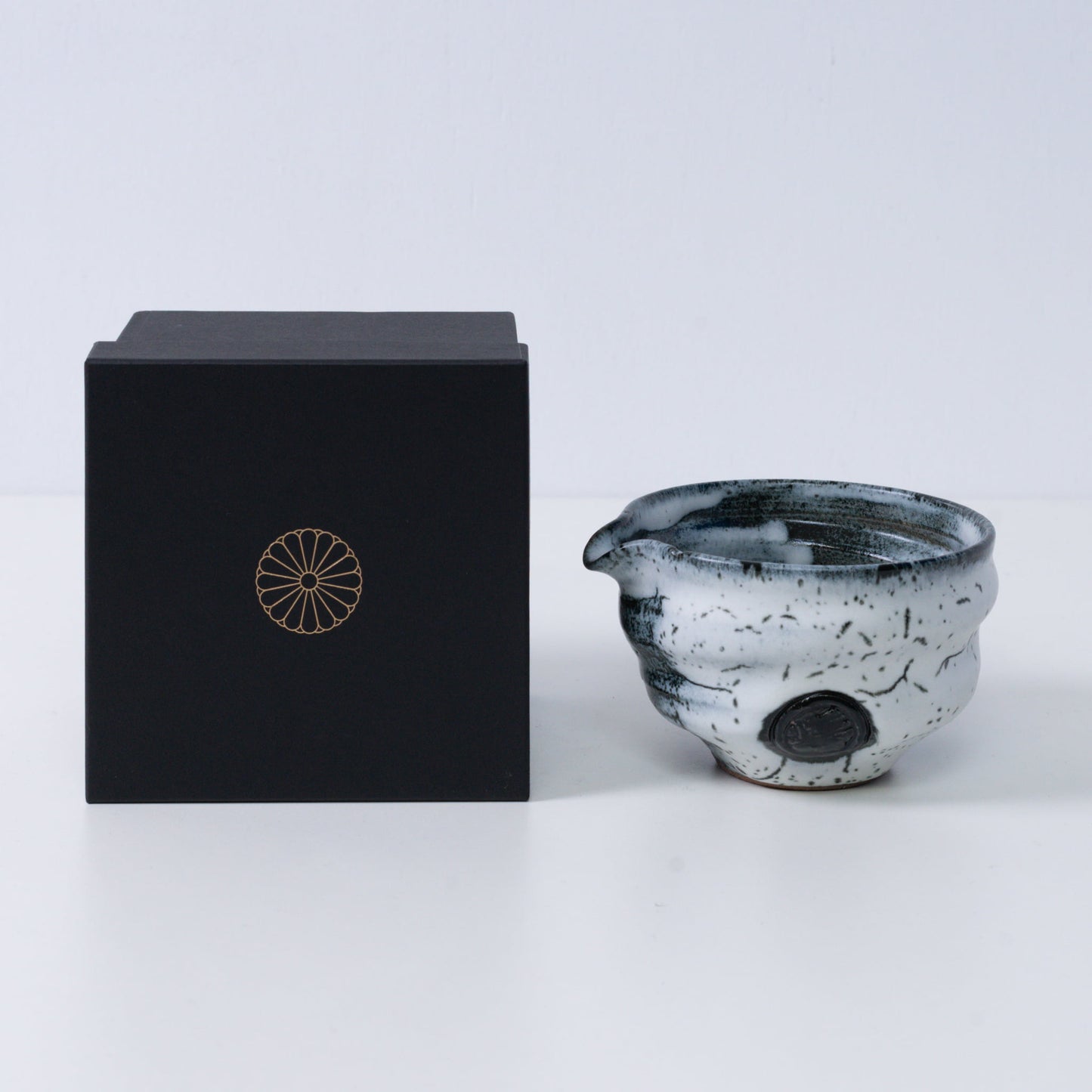 Witte en zwarte Japanse matchakom met tuit "Creative Cloud" aardewerk naast een zwarte geschenkdoos met een gouden chrysantenembleem, uit Japan.