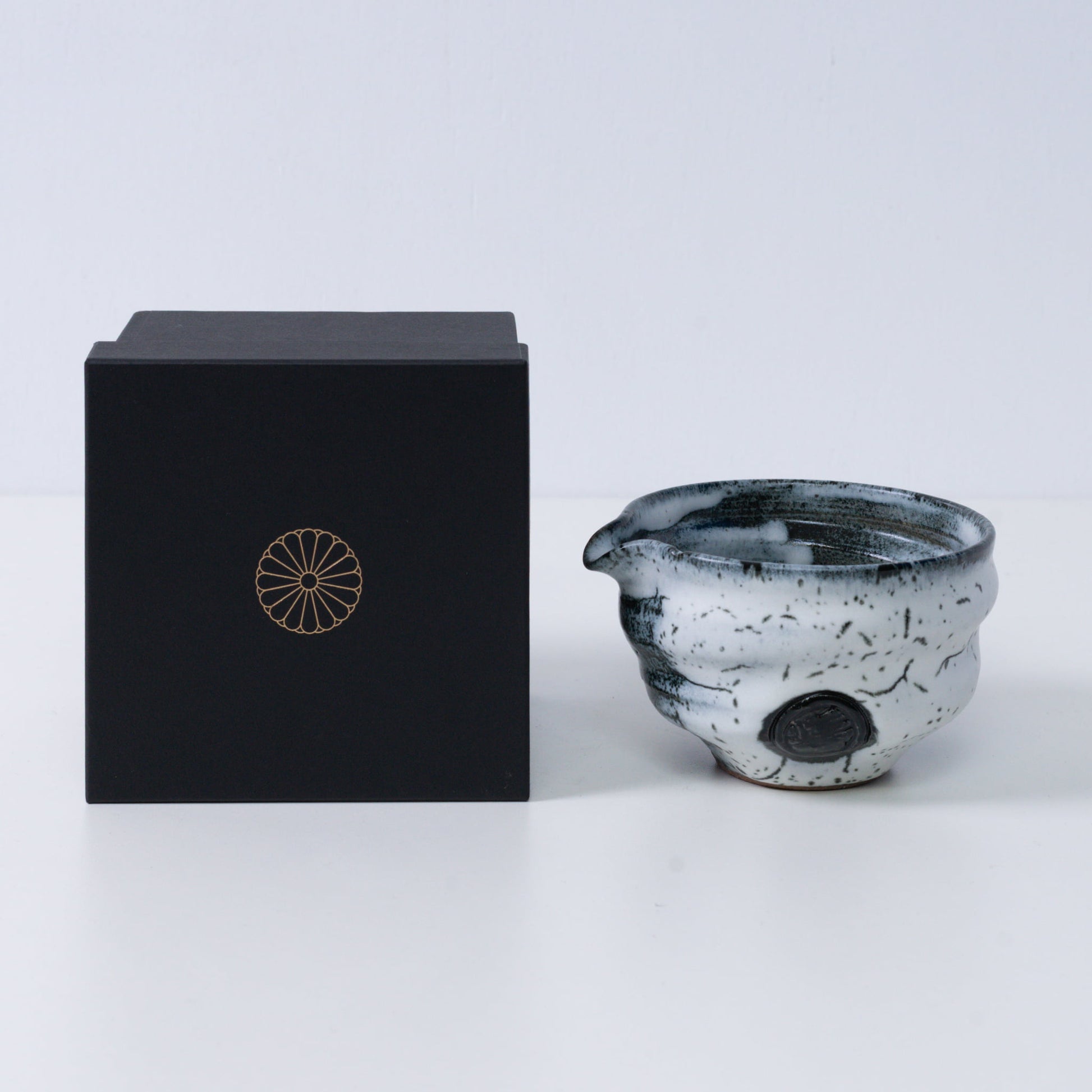 Witte en zwarte Japanse matchakom met tuit "Creative Cloud" aardewerk naast een zwarte geschenkdoos met een gouden chrysantenembleem, uit Japan.