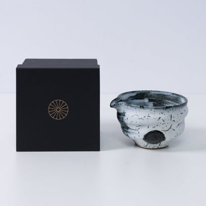 Witte en zwarte Japanse matchakom met tuit "Creative Cloud" aardewerk naast een zwarte geschenkdoos met een gouden chrysantenembleem, uit Japan.