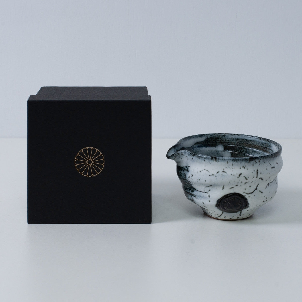 White and black Japanese matcha bowl with spout "Creative Cloud "pottery beside a black gift box with a gold chrysanthemum crest, from Japan.