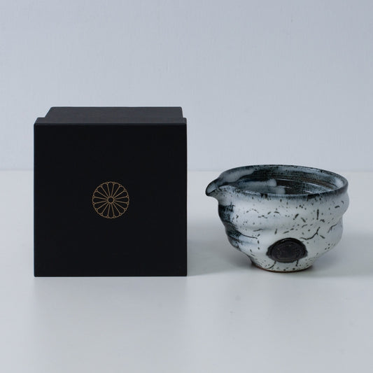 White and black Japanese matcha bowl with spout "Creative Cloud "pottery beside a black gift box with a gold chrysanthemum crest, from Japan.