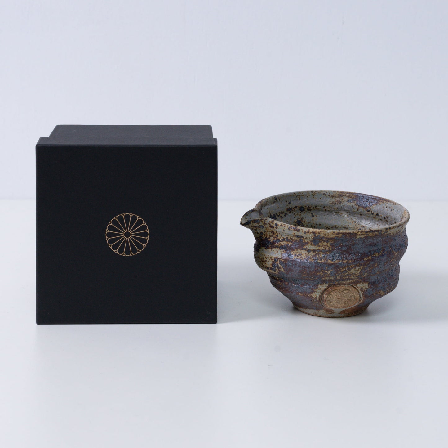 Rustieke bruine Japanse matchakom met tuit "Sakigake" aardewerk naast een zwarte geschenkdoos met een gouden chrysantenembleem, gemaakt in Japan.