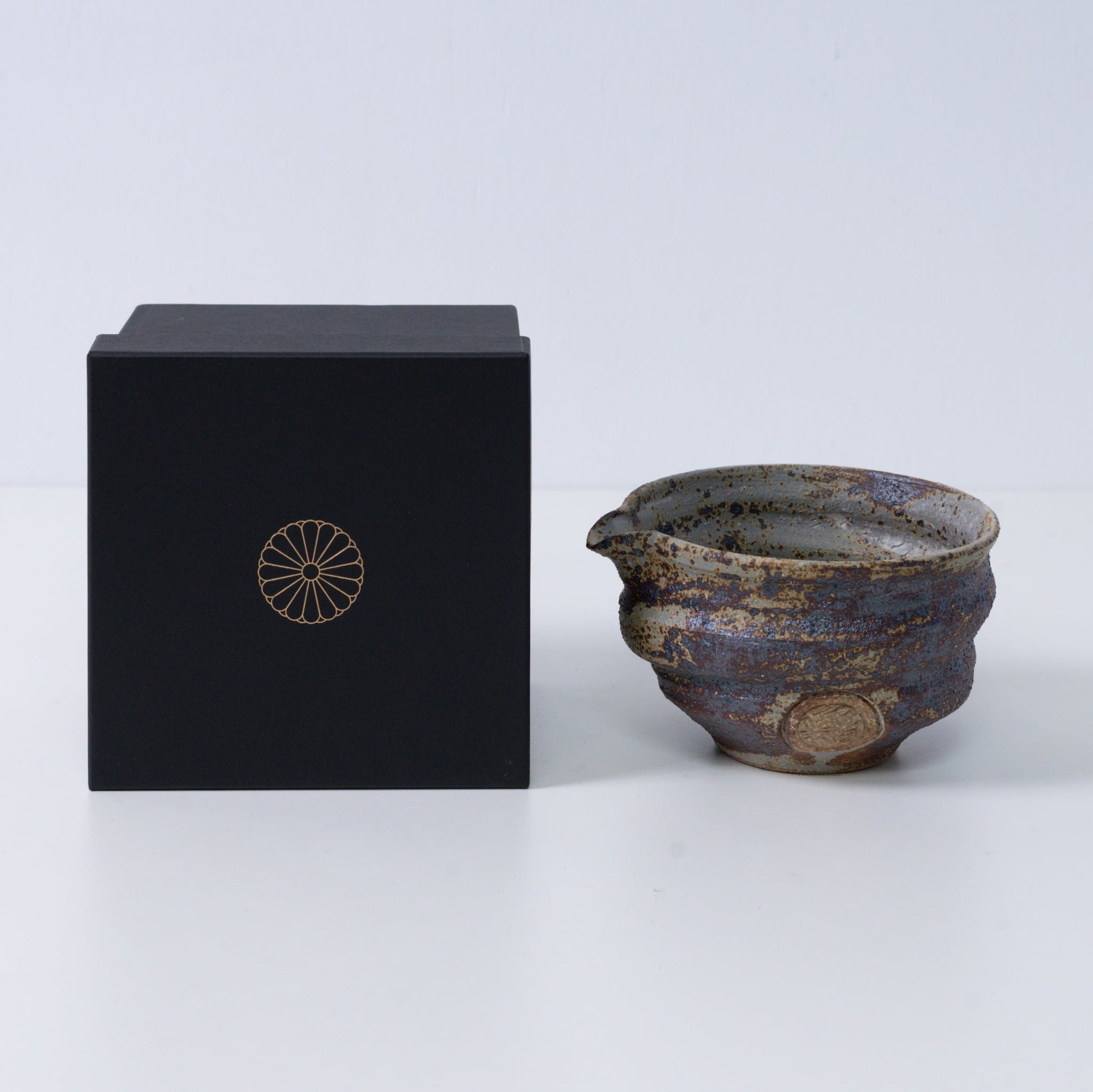 Rustieke bruine Japanse matchakom met tuit "Sakigake" aardewerk naast een zwarte geschenkdoos met een gouden chrysantenembleem, gemaakt in Japan.