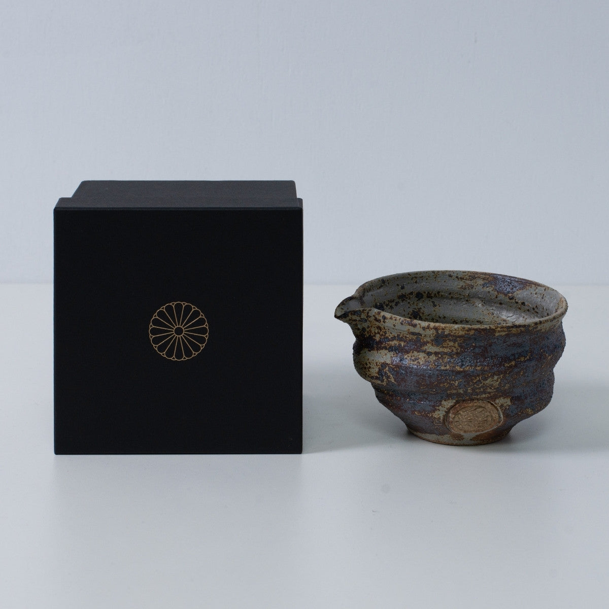 Rustic brown Japanese matcha bowl with spout "Sakigake" pottery next to a black gift box with a gold chrysanthemum crest, made in Japan.