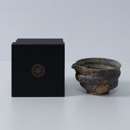 Rustic brown Japanese matcha bowl with spout "Sakigake" pottery next to a black gift box with a gold chrysanthemum crest, made in Japan.